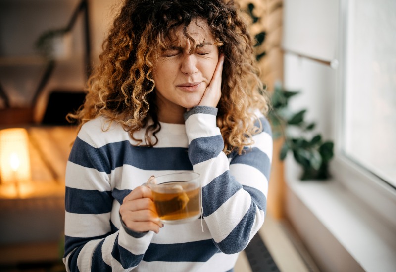 A woman with sensitive teeth after whitening in Brookfield, WI, holding her cheek and a mug of tea